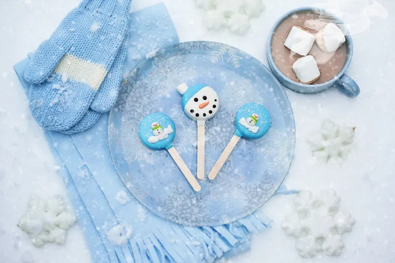 A mug of hot chocolate with marshmallows beside blue knitted mittens and snowflake decorations