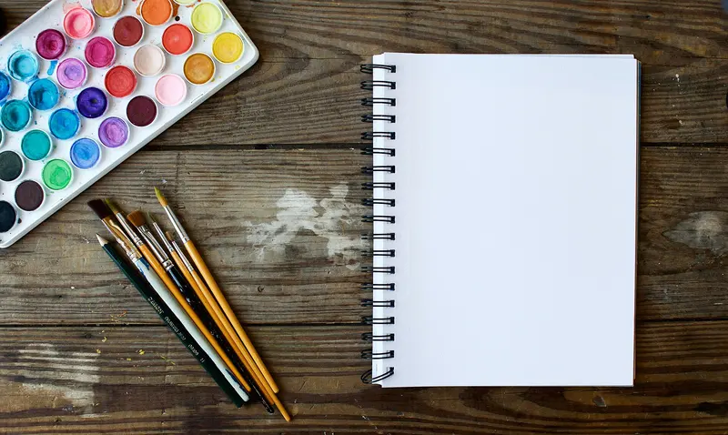 A watercolour paint palette, brushes and a blank spiral sketchbook on a wooden table
