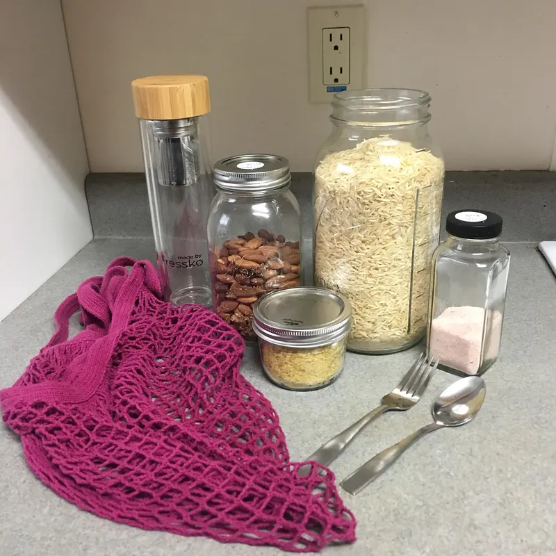 Glass jars of dried food, a reusable bottle, a mesh bag and metal cutlery on a worktop