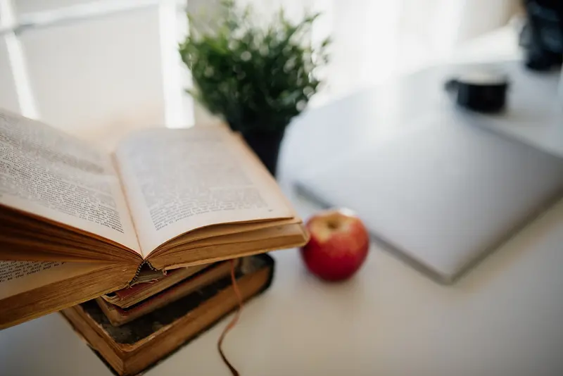 An open book on a pile of books beside a red apple and a pot plant on a bright desk